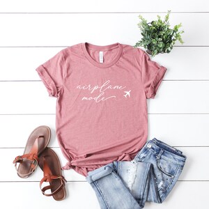 May include: A dusty rose t-shirt with the text "airplane mode" in white script with a white airplane icon. The shirt is laid out on a white wood surface with a pair of brown sandals and blue jeans.