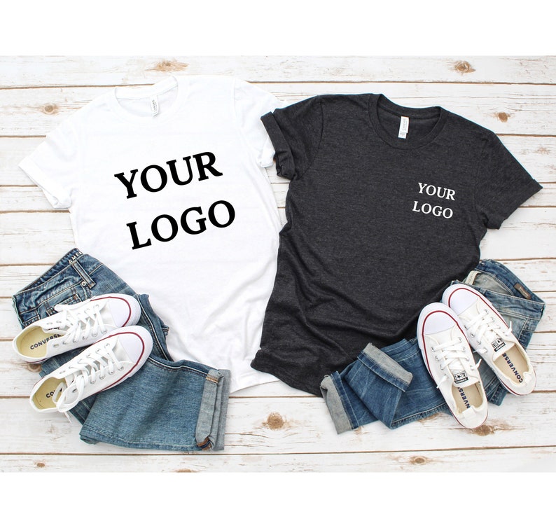 Puede incluir: Dos camisetas, una blanca y otra gris oscuro, ambas con el texto "YOUR LOGO" impreso en ellas. Las camisetas est&aacute;n colocadas sobre una superficie de madera con vaqueros y zapatillas.