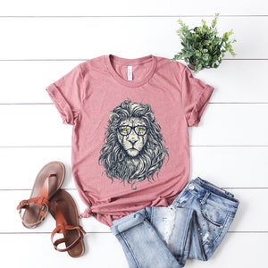 以下が含まれることがあります： 眼鏡をかけたライオンのグラフィックがプリントされたピンクのTシャツ。ライオンは精巧なたてがみと真剣な表情をしています。