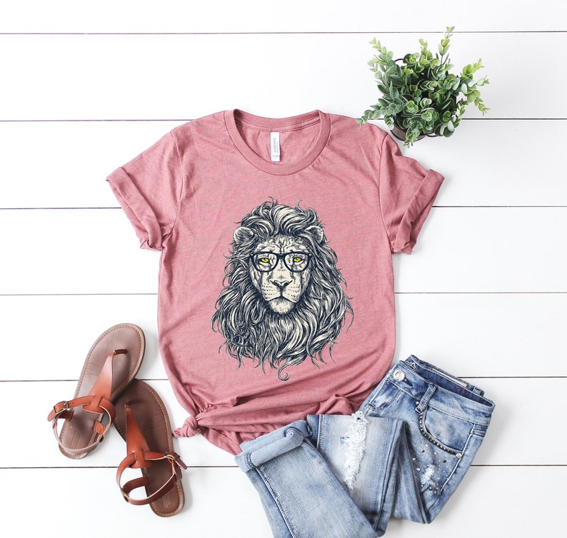 以下が含まれることがあります： 眼鏡をかけたライオンのグラフィックがプリントされたピンクのTシャツ。ライオンは精巧なたてがみと真剣な表情をしています。