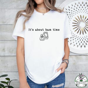 Puede incluir: Camiseta blanca con la frase "it's about bam time" y una ilustraci&oacute;n en blanco y negro de dos fichas de Mahjong. La camiseta es de manga corta y se lleva con vaqueros azules.