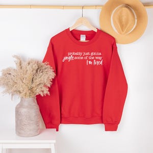 Puede incluir: Una sudadera roja con el texto "probably just gonna jingle some of the way I'm tired" en blanco. Tambi&eacute;n se ven un sombrero de paja y un jarr&oacute;n con flores secas.