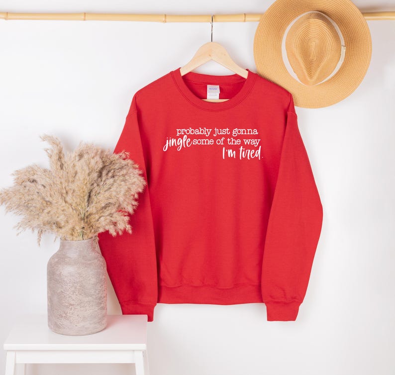 Puede incluir: Una sudadera roja con el texto "probably just gonna jingle some of the way I'm tired" en blanco. Tambi&eacute;n se ven un sombrero de paja y un jarr&oacute;n con flores secas.