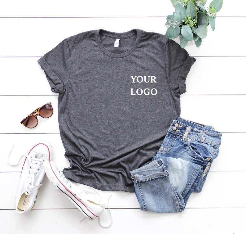 Puede incluir: Una camiseta gris oscuro con el texto "YOUR LOGO" impreso en la parte delantera. La camiseta est&aacute; colocada sobre una superficie de madera blanca con un par de zapatillas blancas, unas gafas de sol y una planta en maceta.