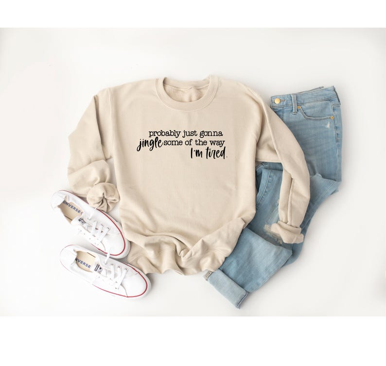 Puede incluir: Una sudadera beige con el texto "probably just gonna jingle some of the way I'm tired" en escritura negra. La sudadera se combina con vaqueros azul claro y zapatillas blancas.