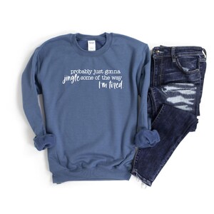 Puede incluir: Una sudadera azul con el texto "probably just gonna jingle some of the way I'm tired" en letras blancas. Vaqueros azul oscuro doblados junto a la sudadera. Los art&iacute;culos est&aacute;n sobre un fondo blanco.