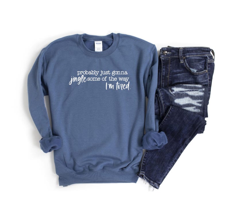 Puede incluir: Una sudadera azul con el texto "probably just gonna jingle some of the way I'm tired" en letras blancas. Vaqueros azul oscuro doblados junto a la sudadera. Los art&iacute;culos est&aacute;n sobre un fondo blanco.
