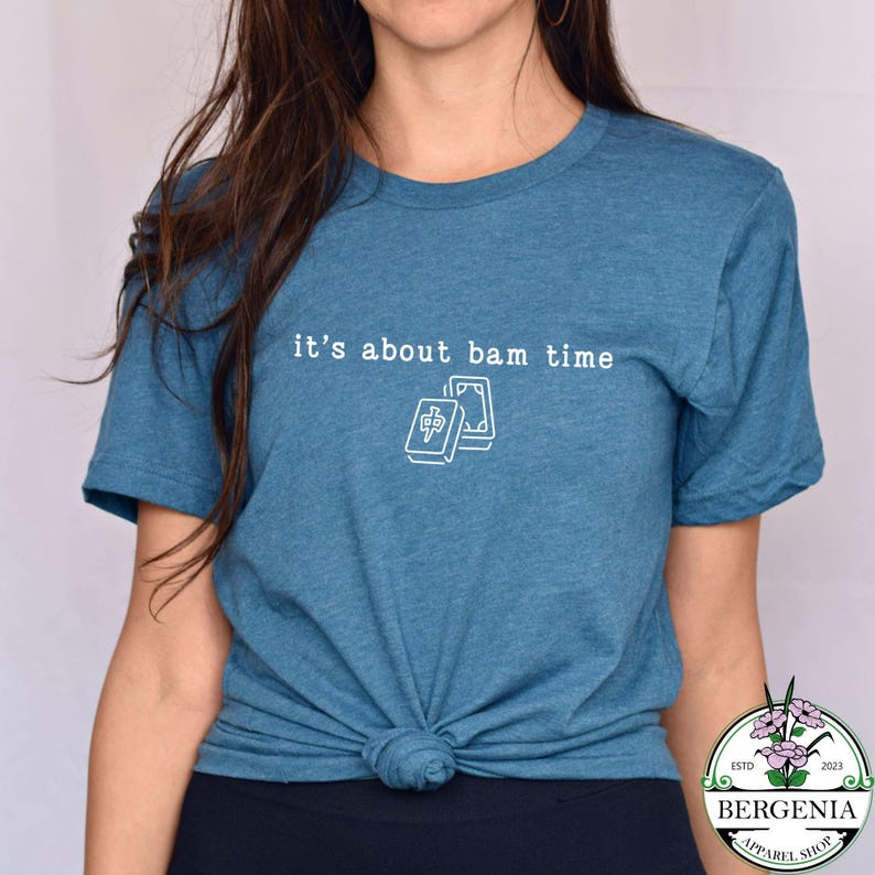 Puede incluir: Camiseta azul turquesa con el texto "it's about bam time" y un gr&aacute;fico blanco de dos fichas de Mahjong. La camiseta tiene mangas cortas y cuello redondo. La parte inferior de la camiseta est&aacute; anudada.