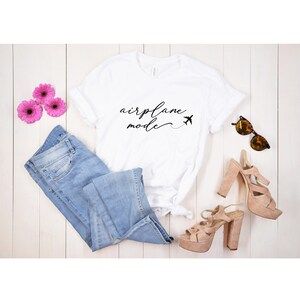May include: White t-shirt with the text "airplane mode" in black script with a small airplane icon. The shirt is laid out on a white wooden surface with blue jeans, pink flowers, sunglasses, and brown heeled sandals.
