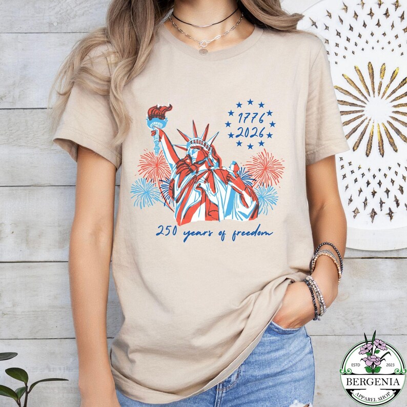 May include: Beige t-shirt featuring a red, white, and blue graphic of the Statue of Liberty with fireworks and the text "1776-2026" and "250 years of freedom". The shirt is a crew neck style.
