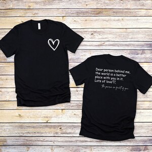 以下が含まれることがあります： 白い文字で書かれた黒いTシャツが2枚。前のTシャツは左胸に小さなハートがあります。後ろのTシャツには「私の後ろにいる人へ、あなたがいる世界はもっと良い場所です。たくさんの愛を込めて、あなたの前にいる人より」と書かれています。