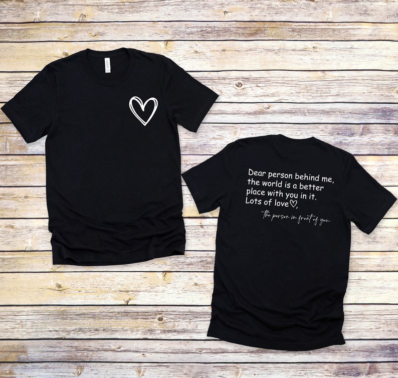 以下が含まれることがあります： 白い文字で書かれた黒いTシャツが2枚。前のTシャツは左胸に小さなハートがあります。後ろのTシャツには「私の後ろにいる人へ、あなたがいる世界はもっと良い場所です。たくさんの愛を込めて、あなたの前にいる人より」と書かれています。