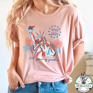 May include: A light pink t-shirt featuring a graphic of the Statue of Liberty in red, white, and blue, with fireworks and the text "1776 2026" and "250 years of freedom". The shirt has rolled-up sleeves.