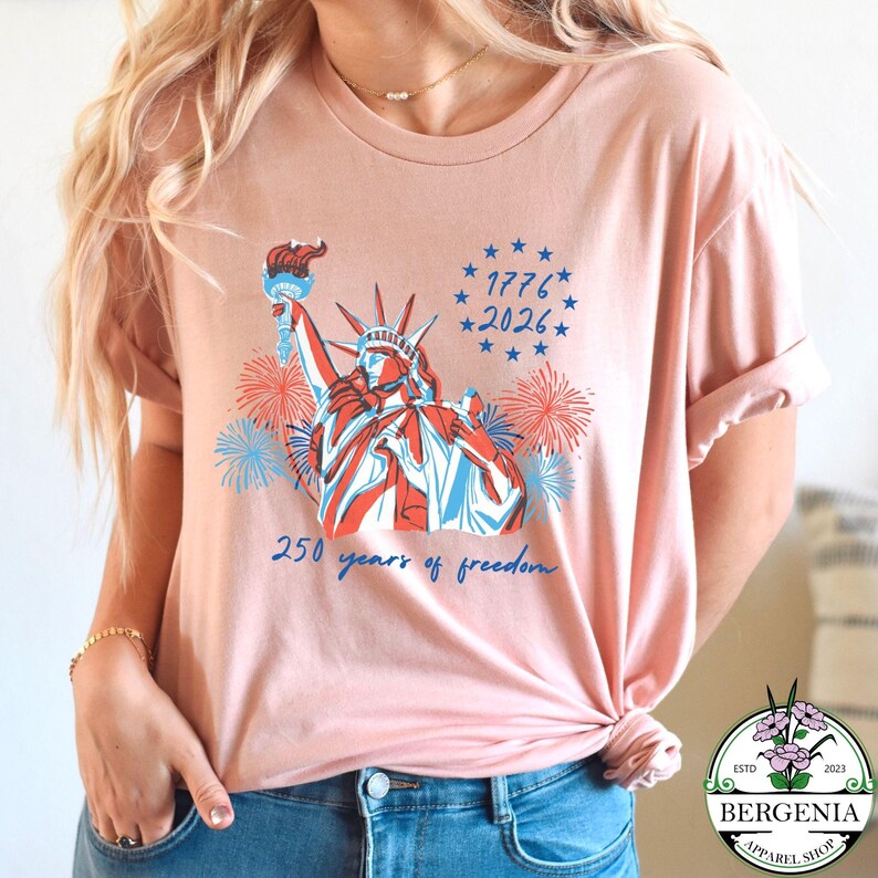 May include: A light pink t-shirt featuring a graphic of the Statue of Liberty in red, white, and blue, with fireworks and the text "1776 2026" and "250 years of freedom". The shirt has rolled-up sleeves.