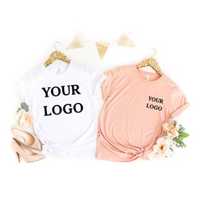 Puede incluir: Dos camisetas, una blanca y otra melocot&oacute;n, colgadas de perchas doradas. Ambas camisetas tienen el texto "YOUR LOGO" impreso.