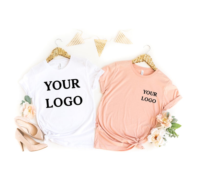Puede incluir: Dos camisetas, una blanca y otra melocot&oacute;n, colgadas de perchas doradas. Ambas camisetas tienen el texto "YOUR LOGO" impreso.