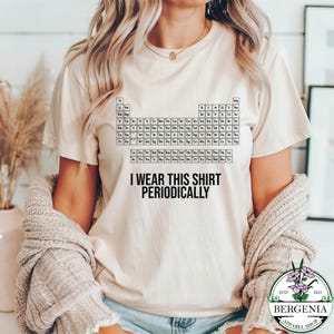 May include: Beige t-shirt featuring a periodic table design and the phrase "I WEAR THIS SHIRT PERIODICALLY" in black text. The shirt is a short-sleeved crew neck style.