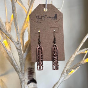 May include: A pair of copper earrings with a tree design. The earrings are hanging from a brown card with the text "Trent Earrings" printed on it.