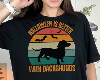 May include: Black t-shirt with a Halloween-themed graphic. The design features a dachshund silhouette, bats, and the text "Halloween is Better With Dachshunds" in orange and white against a sunset-style background.