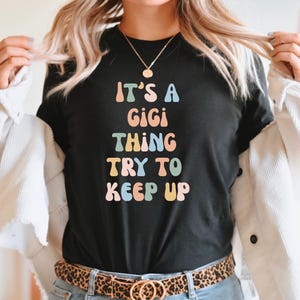 Może przedstawiać: Czarny T-shirt z napisem "It's a Gigi thing try to keep up" w kolorowych, retro literach.