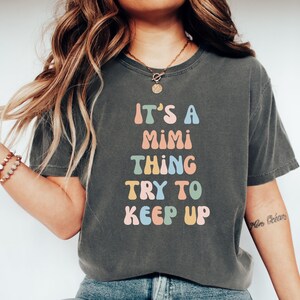 Może przedstawiać: Szary t-shirt z napisem "It's a Mimi thing try to keep up" w kolorowych, stylizowanych literach.