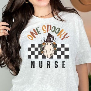 Może przedstawiać: Jasnoszara koszulka z napisem "ONE SPOOKY NURSE" w kolorze pomarańczowym, brązowym i czarnym. Projekt przedstawia ducha w kapeluszu czarownicy na czarno-białym szachownicowym tle z kwiatowymi akcentami.