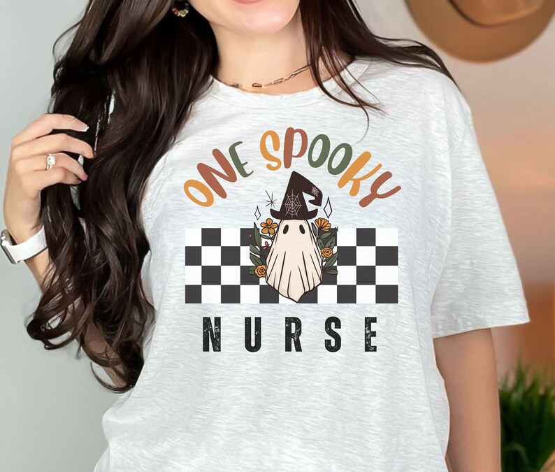 Może przedstawiać: Jasnoszara koszulka z napisem "ONE SPOOKY NURSE" w kolorze pomarańczowym, brązowym i czarnym. Projekt przedstawia ducha w kapeluszu czarownicy na czarno-białym szachownicowym tle z kwiatowymi akcentami.