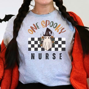 Może przedstawiać: Jasnoszara koszulka z napisem "ONE SPOOKY NURSE" w kolorze pomarańczowym i czarnym. Koszulka przedstawia ducha w kapeluszu czarownicy na czarno-białym szachownicowym tle z kwiatowymi akcentami. Motyw Halloween.