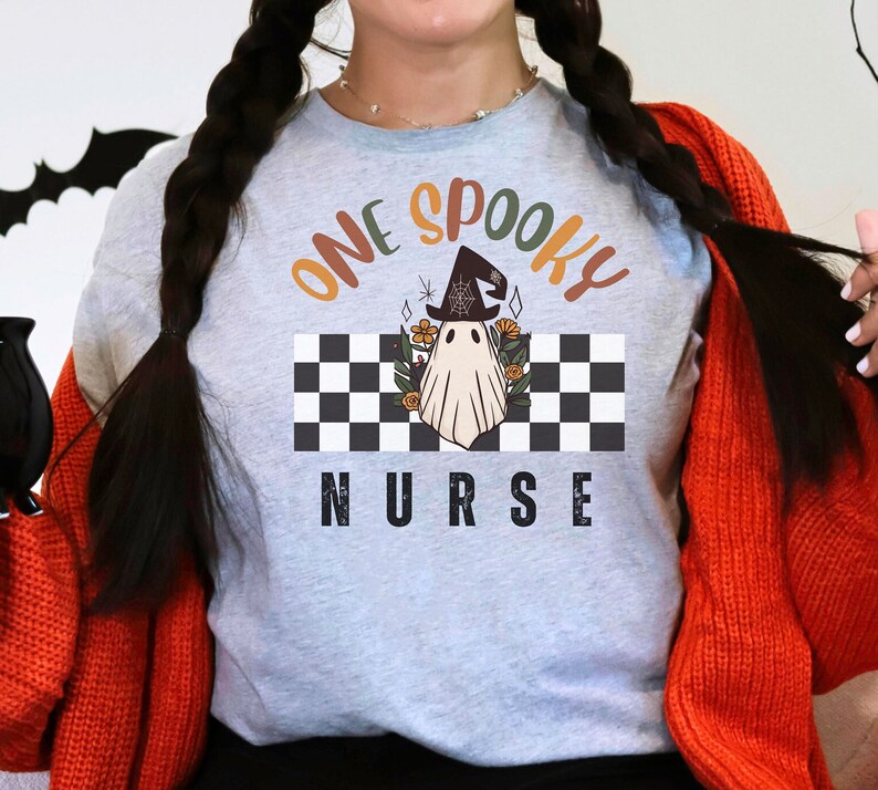 Może przedstawiać: Jasnoszara koszulka z napisem "ONE SPOOKY NURSE" w kolorze pomarańczowym i czarnym. Koszulka przedstawia ducha w kapeluszu czarownicy na czarno-białym szachownicowym tle z kwiatowymi akcentami. Motyw Halloween.