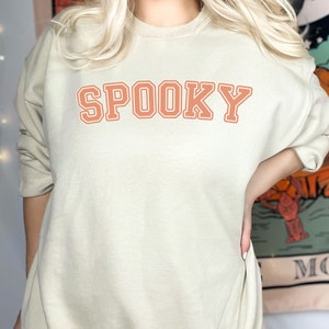 Może przedstawiać: Beżowa bluza z napisem "SPOOKY" w pomarańczowych literach. Litery mają białą obwódkę. Bluza jest w neutralnym kolorze i ma długie rękawy.
