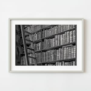 May include: A black and white framed wall art print depicting a tall wooden library ladder leaning against floor-to-ceiling bookshelves filled with numerous antique, leather-bound books arranged in neat rows across multiple wooden shelves.