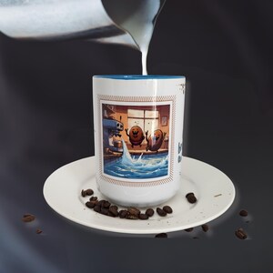 May include: White ceramic coffee mug with a blue interior rim, featuring a cartoon illustration of two smiling coffee beans in a pool. The mug sits on a white saucer with scattered coffee beans, with milk being poured into the mug.