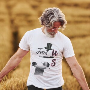 Peut inclure: T-shirt blanc avec l'inscription "Just us" et un dessin de chapeau haut de forme. Le t-shirt présente une image stylisée de deux personnages avec des chapeaux. L'image est placée sur un fond de champ de blé flou.