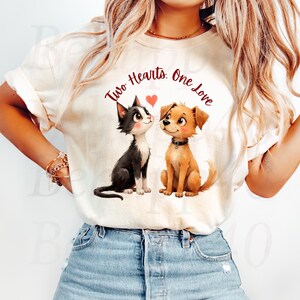 Peut inclure: T-shirt crème avec un chat et un chien de dessin animé, le texte "Two Hearts, One Love" et un cœur. Le chat est noir et blanc, le chien est beige. Le t-shirt est de style décontracté.