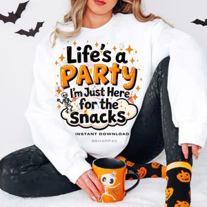 Peut inclure: Sweat-shirt blanc avec l'inscription "Life's a PARTY I'm Just Here for the Snacks" en orange et noir. L'image comprend une tasse orange sur le thème d'Halloween avec un motif de squelette et des leggings noirs. Des découpes de chauves-souris noires sont en arrière-plan.