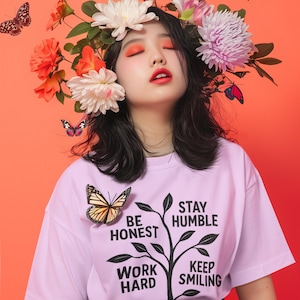 Peut inclure: Une personne portant un t-shirt violet clair avec un papillon et les mots "BE HONEST, WORK HARD, STAY HUMBLE, KEEP SMILING." La personne porte une couronne de fleurs et des papillons.