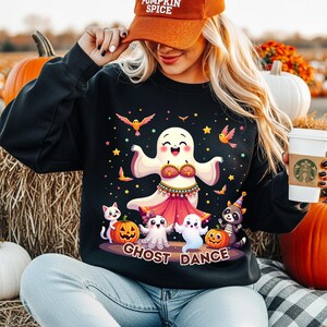 Peut inclure: Sweat-shirt noir avec un motif coloré sur le thème d'Halloween. Le motif représente un fantôme dansant, des citrouilles, des étoiles et le texte "Ghost Dance". Le sweat-shirt est porté avec une casquette "Pumpkin Spice".