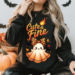 Puede incluir: Sudadera negra con el texto "Cute & Fine" en naranja y amarillo. El diseño presenta un fantasma de dibujos animados con un lazo con estampado de leopardo, rodeado de murciélagos y hojas de otoño. Una calabaza es visible en el fondo.
