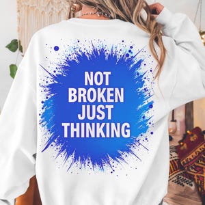 Peut inclure: Sweat-shirt blanc avec un motif bleu au dos. Le motif présente les mots "NOT BROKEN JUST THINKING" en blanc, sur un fond bleu éclaboussé. Le sweat-shirt est de style décontracté et confortable.