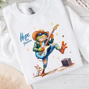May include: White sweatshirt featuring a whimsical illustration of a frog playing an electric guitar, wearing a cowboy hat and sunglasses. The design includes the text "Hope for Tomorrow" and a small cat. A small amp is also included in the design.
