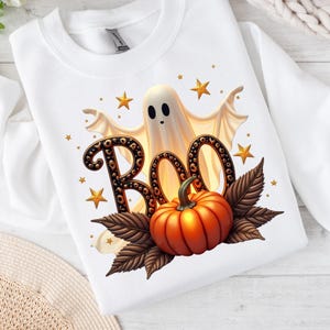 May include: White sweatshirt with a Halloween-themed graphic. The design features a friendly ghost, the word "BOO" in a decorative font, a bright orange pumpkin, and gold stars. The overall theme is festive and seasonal.