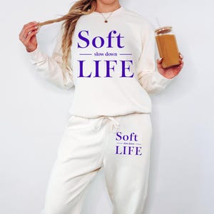 A woman wearing a white sweatshirt with the text "Soft slow down life" printed on it, holding a glass of what appears to be a beverage.