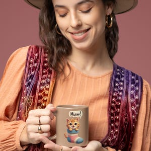 Peut inclure: Mug en céramique beige avec un chat de dessin animé sortant d'une chaussette rayée. Le chat est orange et marron, avec le mot "Miaw!" dans une bulle. La tasse a une anse blanche.