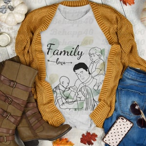 Peut inclure: Composition à plat avec un t-shirt blanc orné d'une illustration familiale et des mots "Family love". Il est associé à un cardigan moutarde, des bottes marron et un jean bleu. Des feuilles d'automne et des citrouilles complètent la scène.
