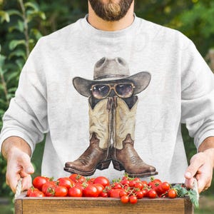 Peut inclure: Un sweat-shirt gris clair avec une illustration à l'aquarelle de bottes de cowboy, d'un chapeau et de lunettes de soleil. Les bottes sont marron et beige, le chapeau est gris et les lunettes de soleil ont une monture dorée. Le sweat-shirt est tenu par une personne.