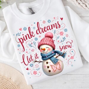Peut inclure: Sweat-shirt blanc avec un motif de bonhomme de neige. Le bonhomme de neige porte un bonnet rose et une écharpe bleue, avec le texte "pink dreams let it snow". Des coeurs et des fleurs décorent le motif.