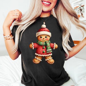 Peut inclure: T-shirt noir avec une illustration de bonhomme en pain d'épices. Le bonhomme en pain d'épices porte un bonnet, une écharpe et un pull rouges avec des détails verts. Le design est sur le thème des fêtes.