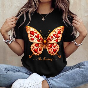 Peut inclure: T-shirt noir avec un motif de papillon. Le papillon a des motifs en forme de cœur rouge sur ses ailes et des accents dorés. Les mots "Be Loving" sont écrits en dessous dans une police script. Le t-shirt est à col rond.