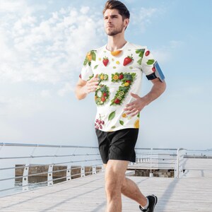 Peut inclure: T-shirt blanc orné d'un motif de fruits et du chiffre "67" composé de fruits. Le motif comprend des fraises, des oranges, des raisins et des kiwis. La personne porte un short noir et court sur une jetée en bois.