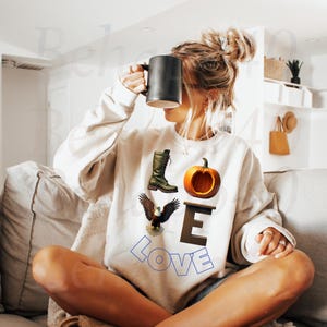 Peut inclure: Sweat-shirt crème avec un motif graphique représentant une botte verte, une citrouille et un aigle. Le mot "LOVE" est épelé avec les lettres et les images. Une tasse noire est tenue devant le visage.